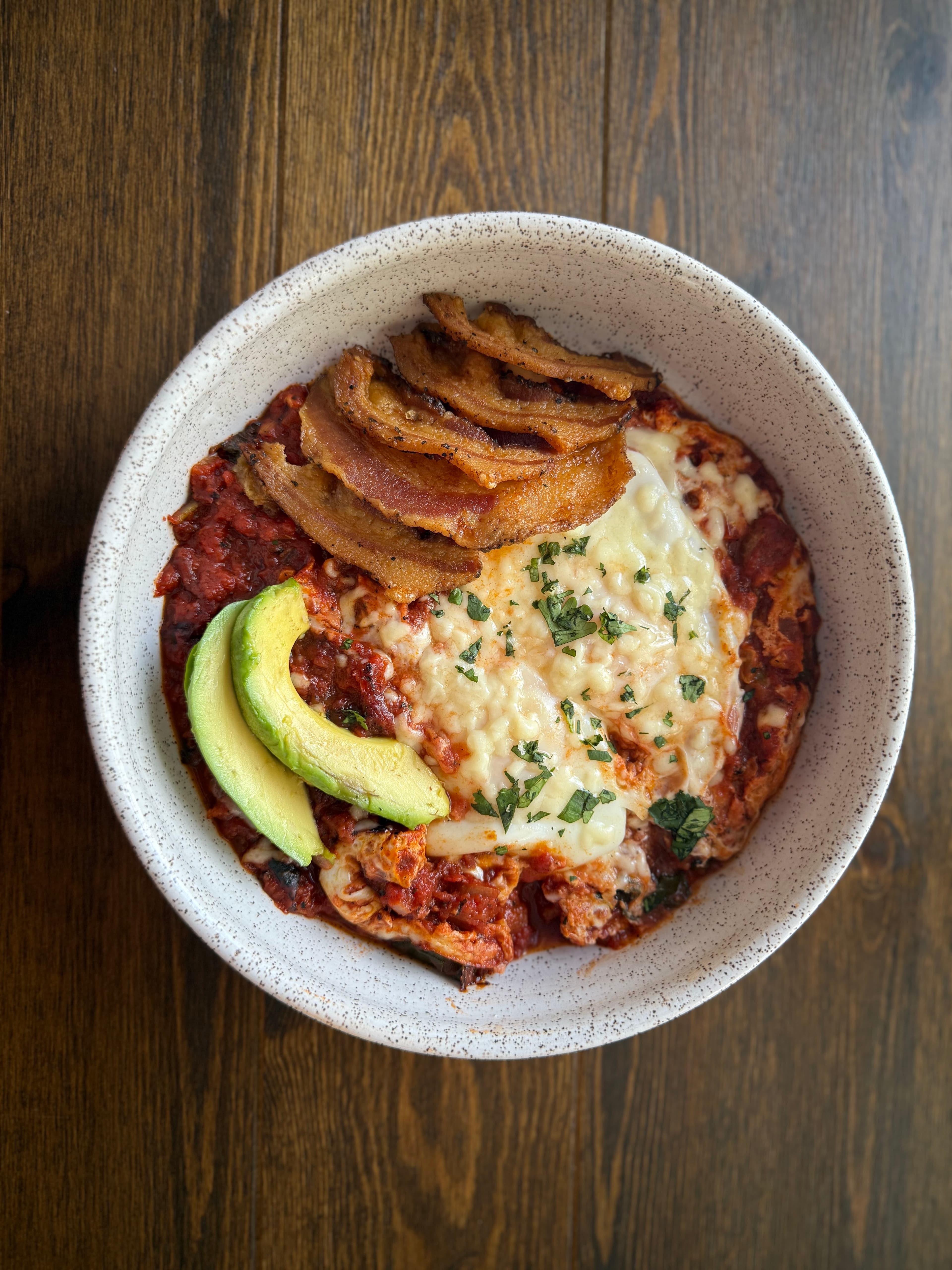 A bowl of baked dish topped with melted cheese, avocado slices, and crispy meat.