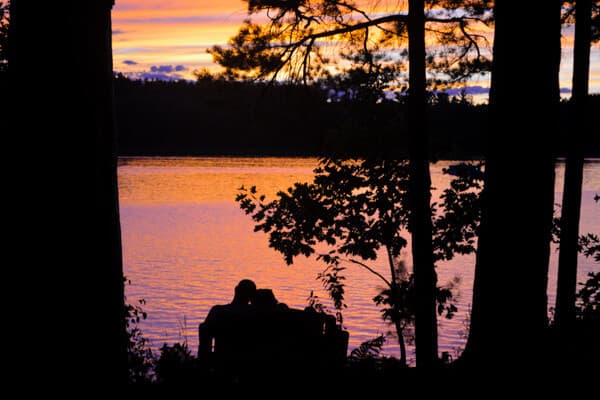 Silhouette of a couple seated by a lake at sunset, surrounded by trees.