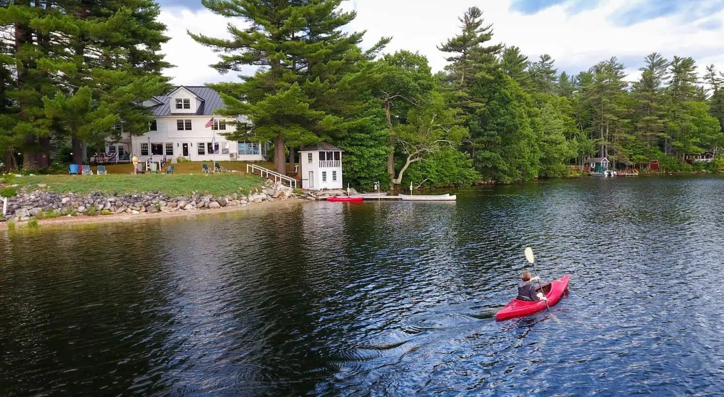 A person paddles a red kayak on a calm lake near a waterfront house surrounded by trees.