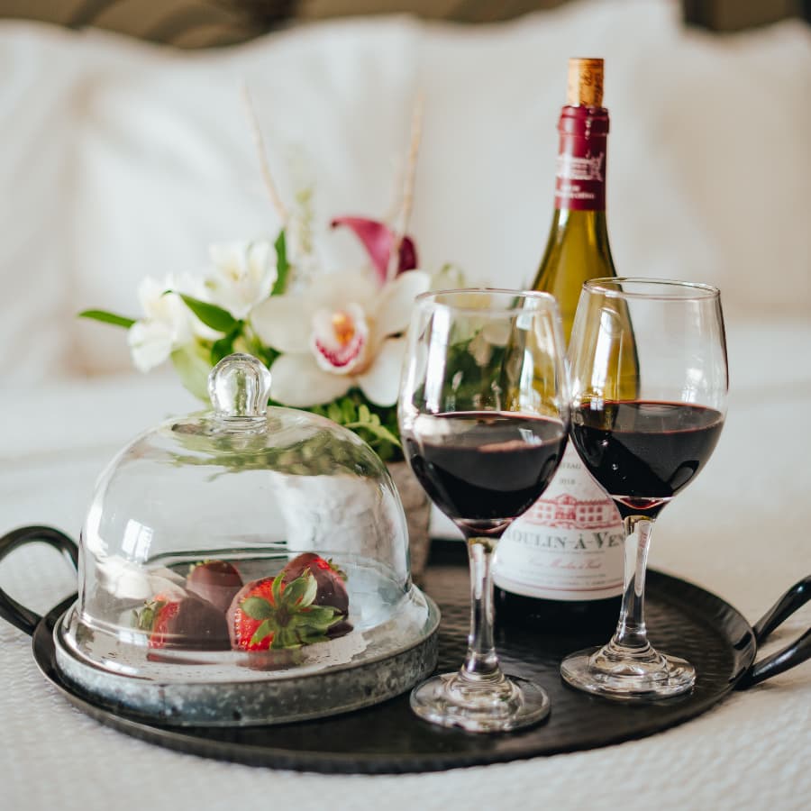 A tray with two glasses of red wine, chocolate-covered strawberries under a cloche, and a bottle of wine, accompanied by a floral arrangement.