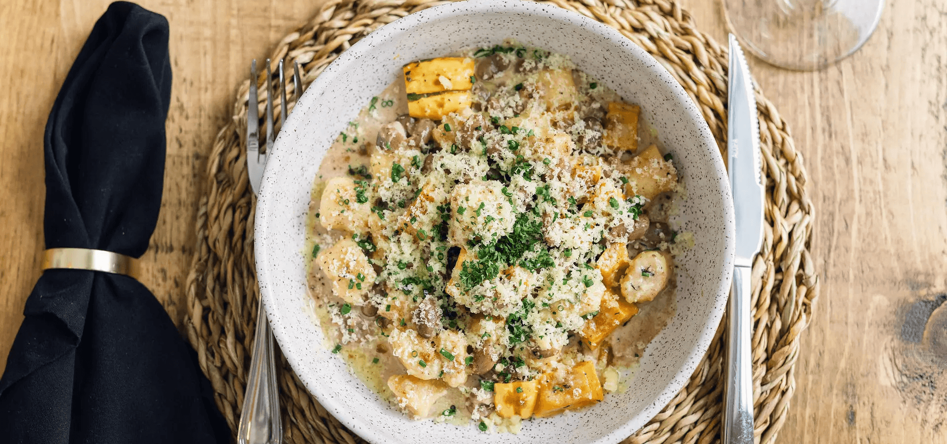 A bowl of creamy pasta topped with herbs and cheese, placed on a woven placemat with a black napkin and cutlery.