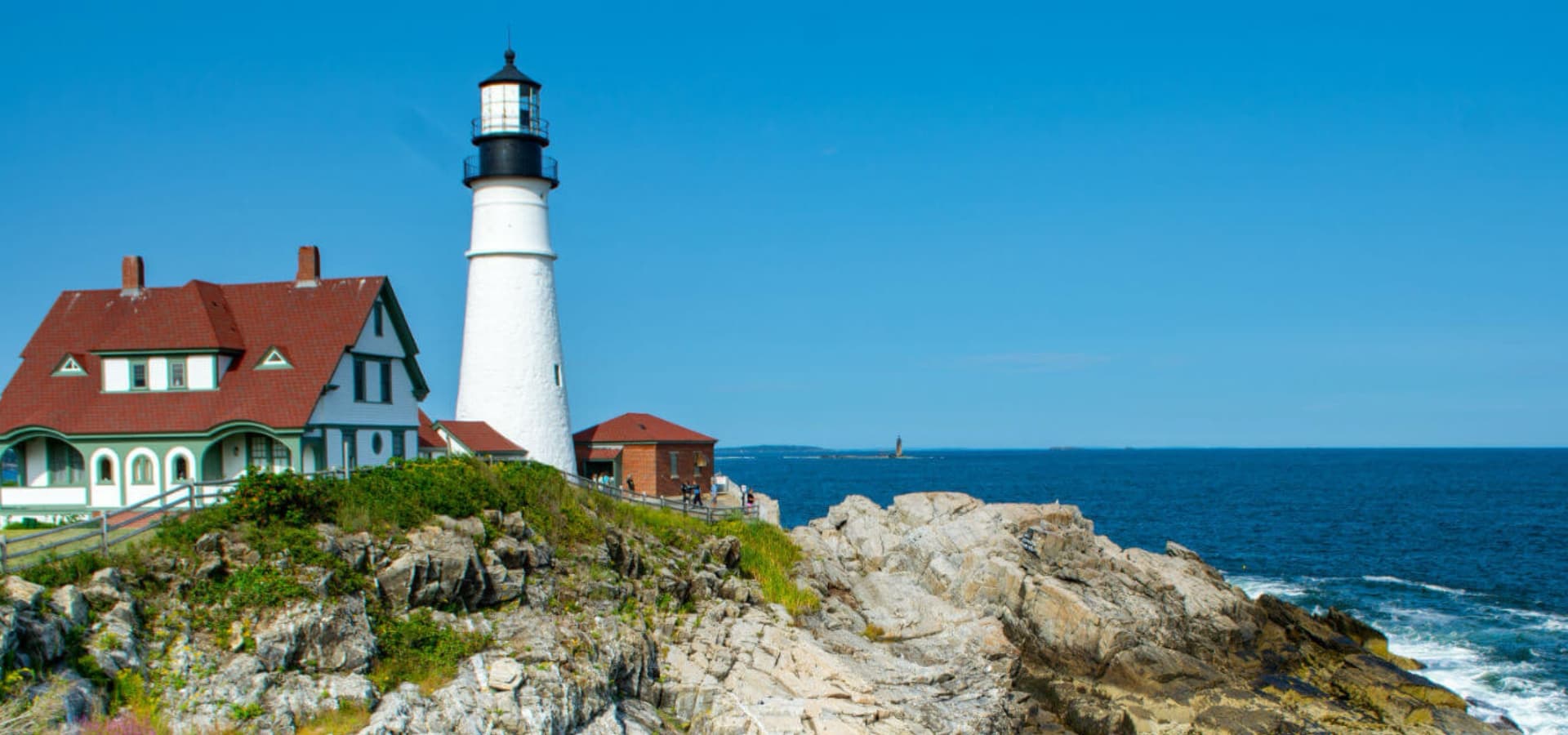A white lighthouse next to a red-roofed house overlooks rocky shores and a blue ocean.