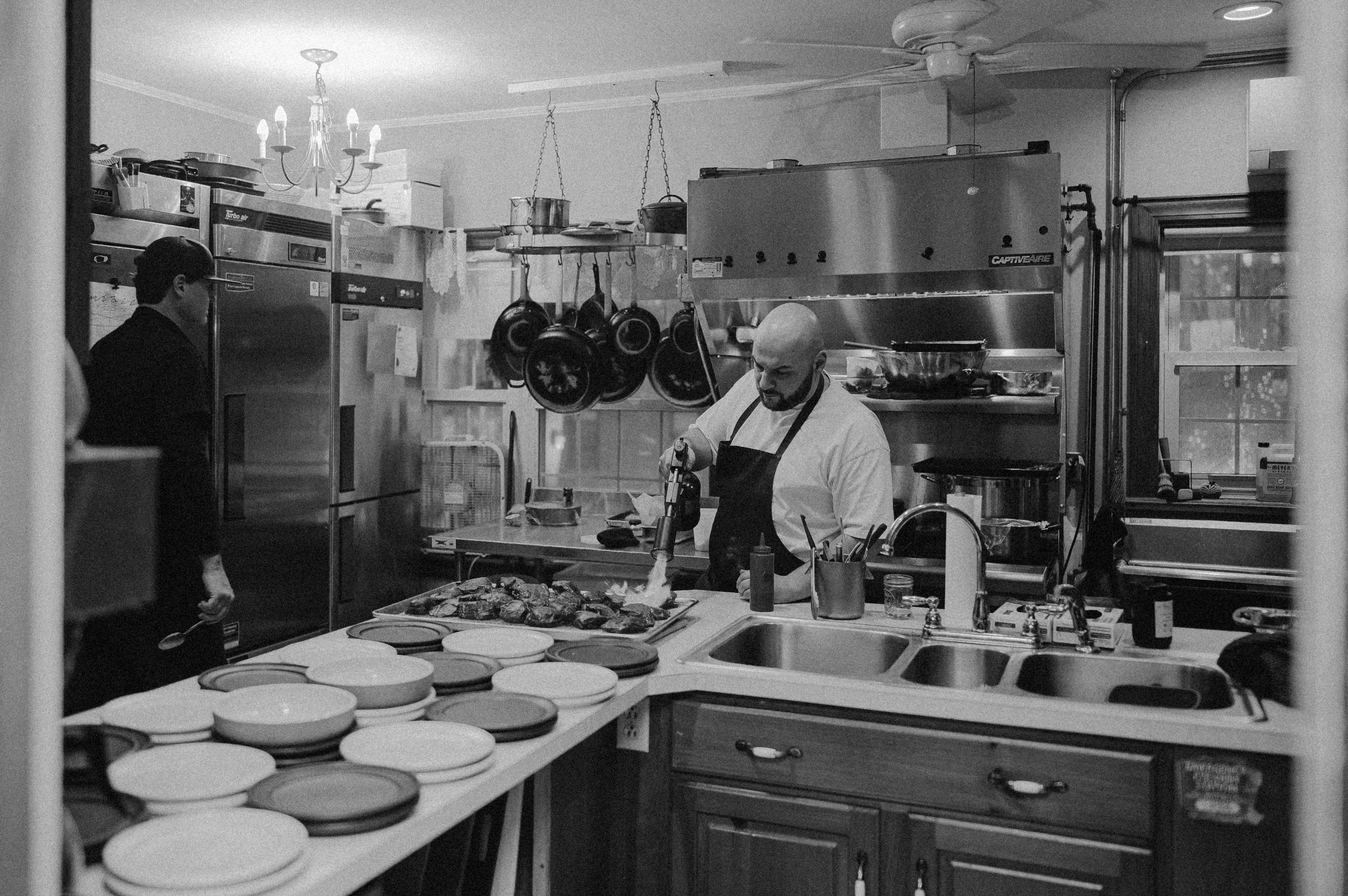 A chef uses a torch to caramelize food in a busy kitchen.