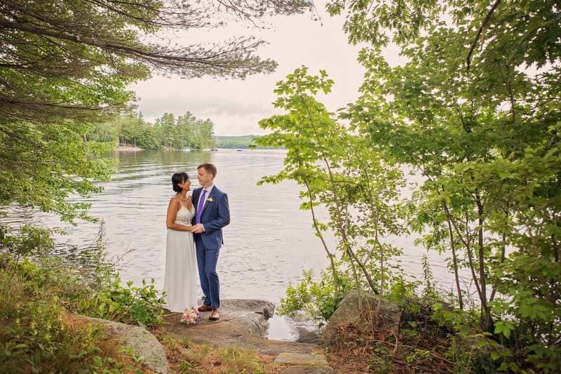 A bride and groom stand together by a tranquil lake, surrounded by lush greenery.