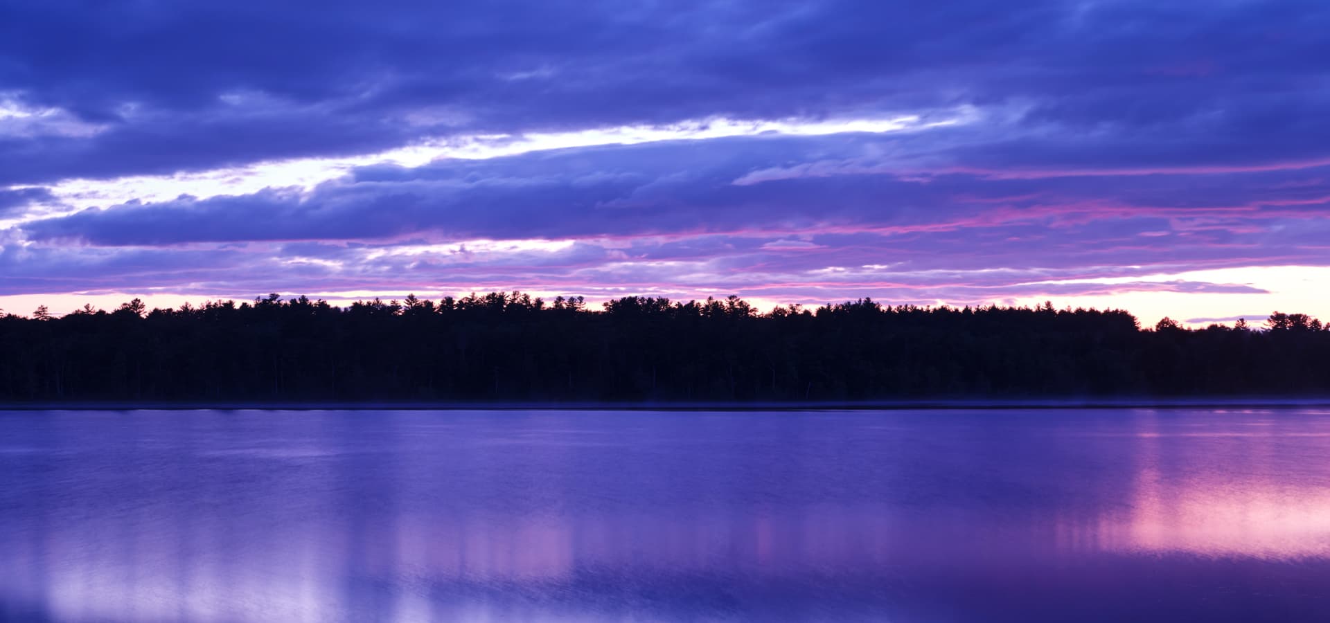 A tranquil lake reflects vibrant purple and blue hues of a sunset, framed by a tree line.
