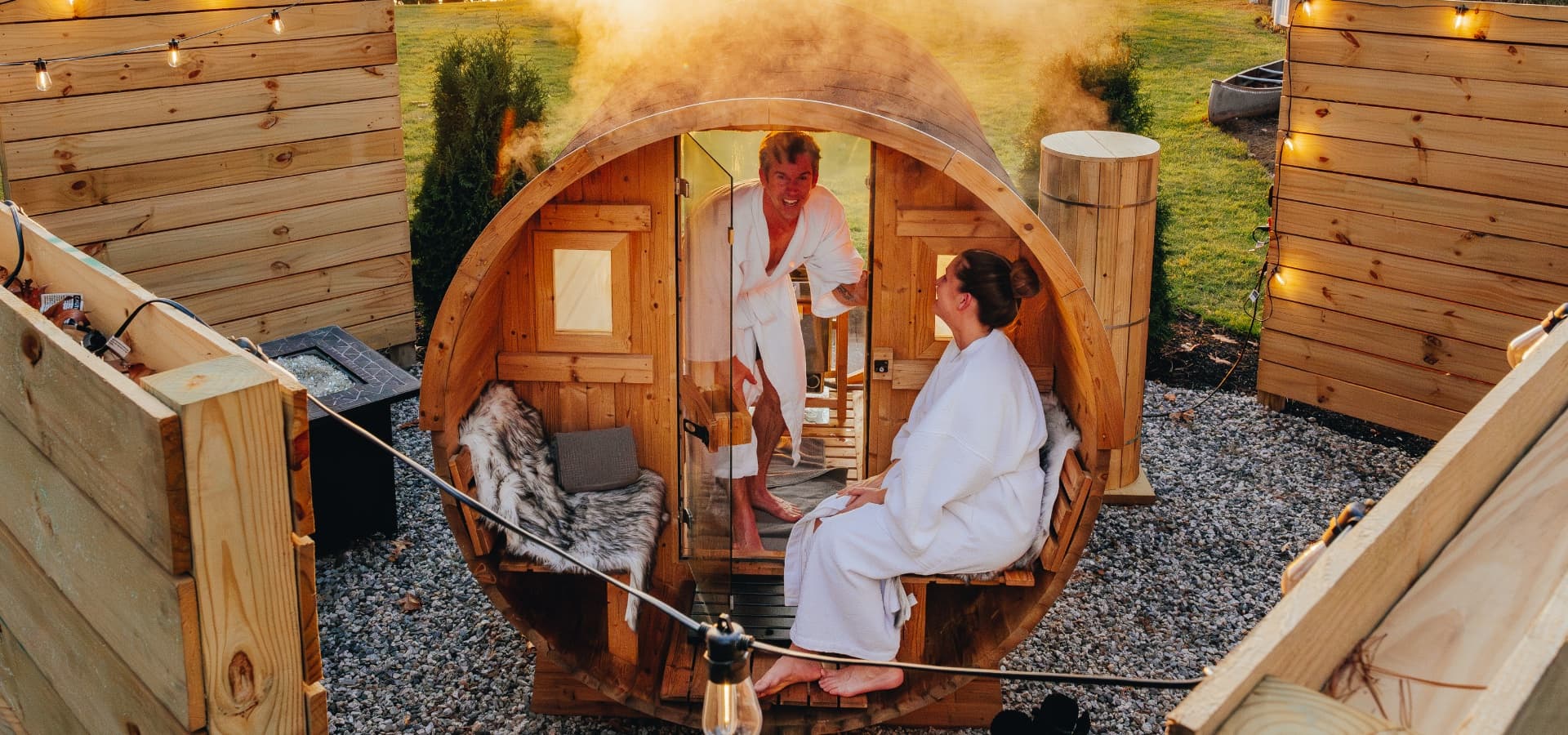 A man and a woman in bathrobes enjoy a sauna experience surrounded by wooden structures and greenery.