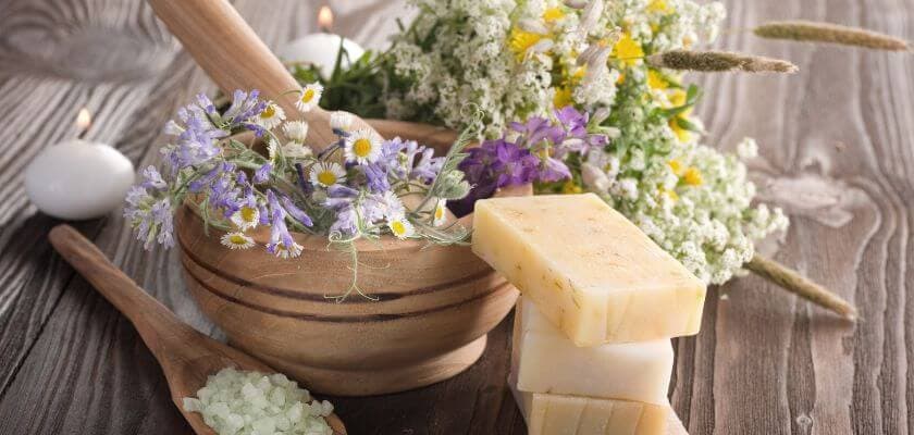 A wooden mortar filled with fresh flowers sits beside soap bars and a wooden spoon with green salt on a rustic table.