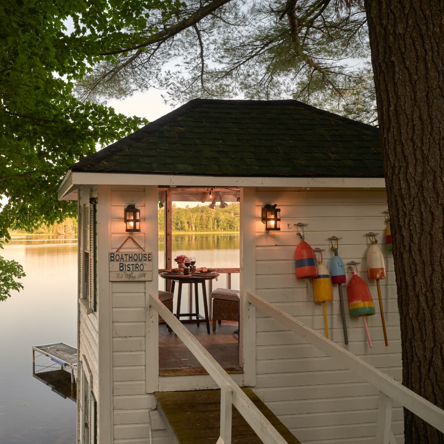 A charming boathouse bistro overlooking a tranquil lake, with a table set for dining and colorful buoys hanging nearby.