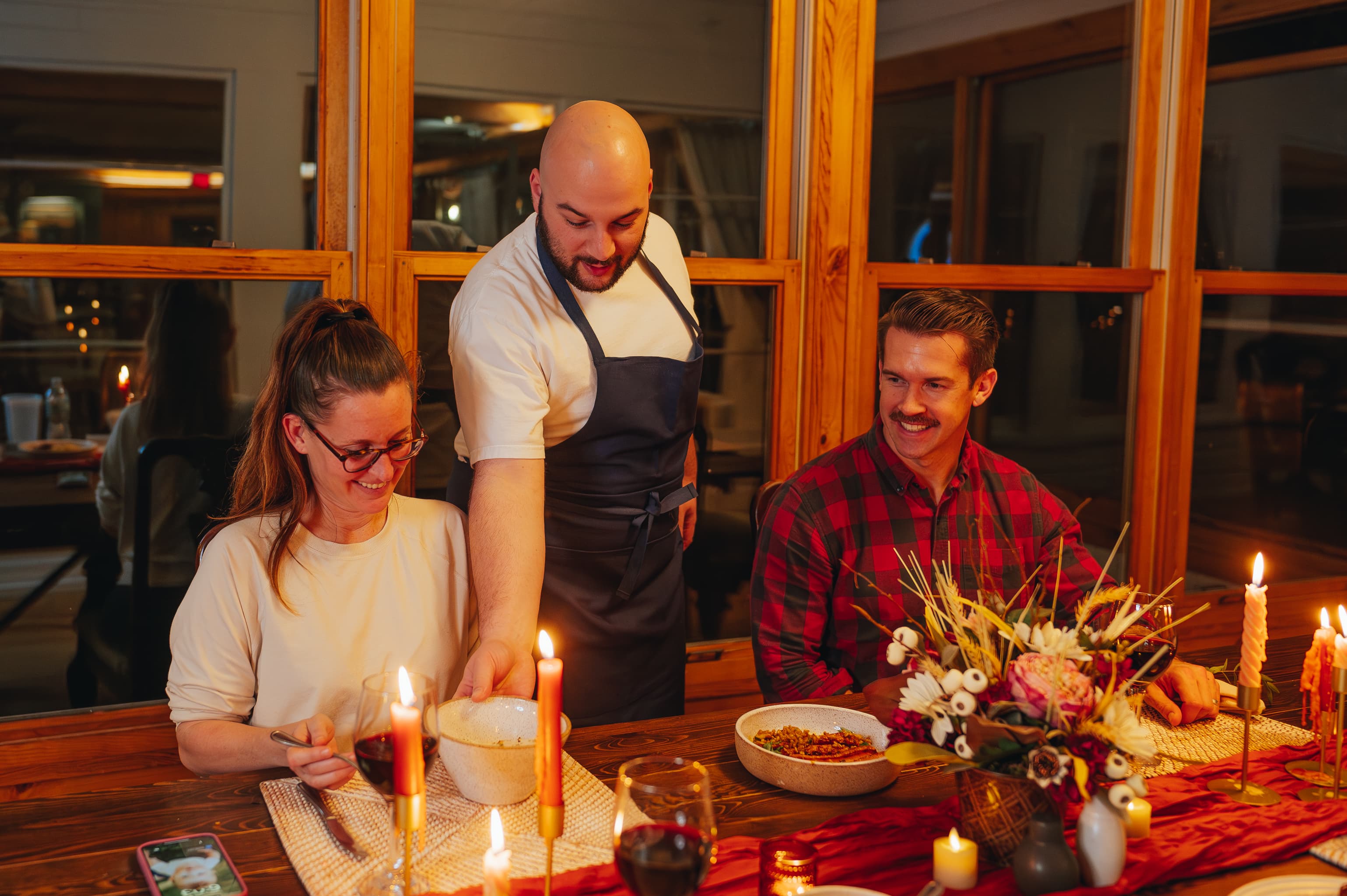 A man in an apron serves food to two smiling diners at a candlelit table.