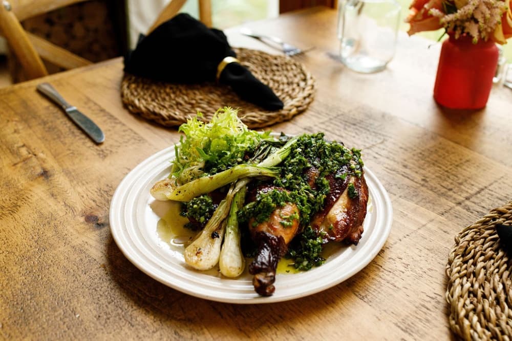 A plate of roasted chicken garnished with green herbs, served with grilled leeks and a side of lettuce, on a rustic wooden table.