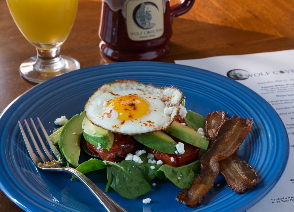 A blue plate with a salad topped with a fried egg, avocado, bacon, and feta cheese, accompanied by a glass of orange juice and a mug. A blue plate with a salad topped with a fried egg, avocado, bacon, and feta cheese, accompanied by a glass of orange juice and a mug.