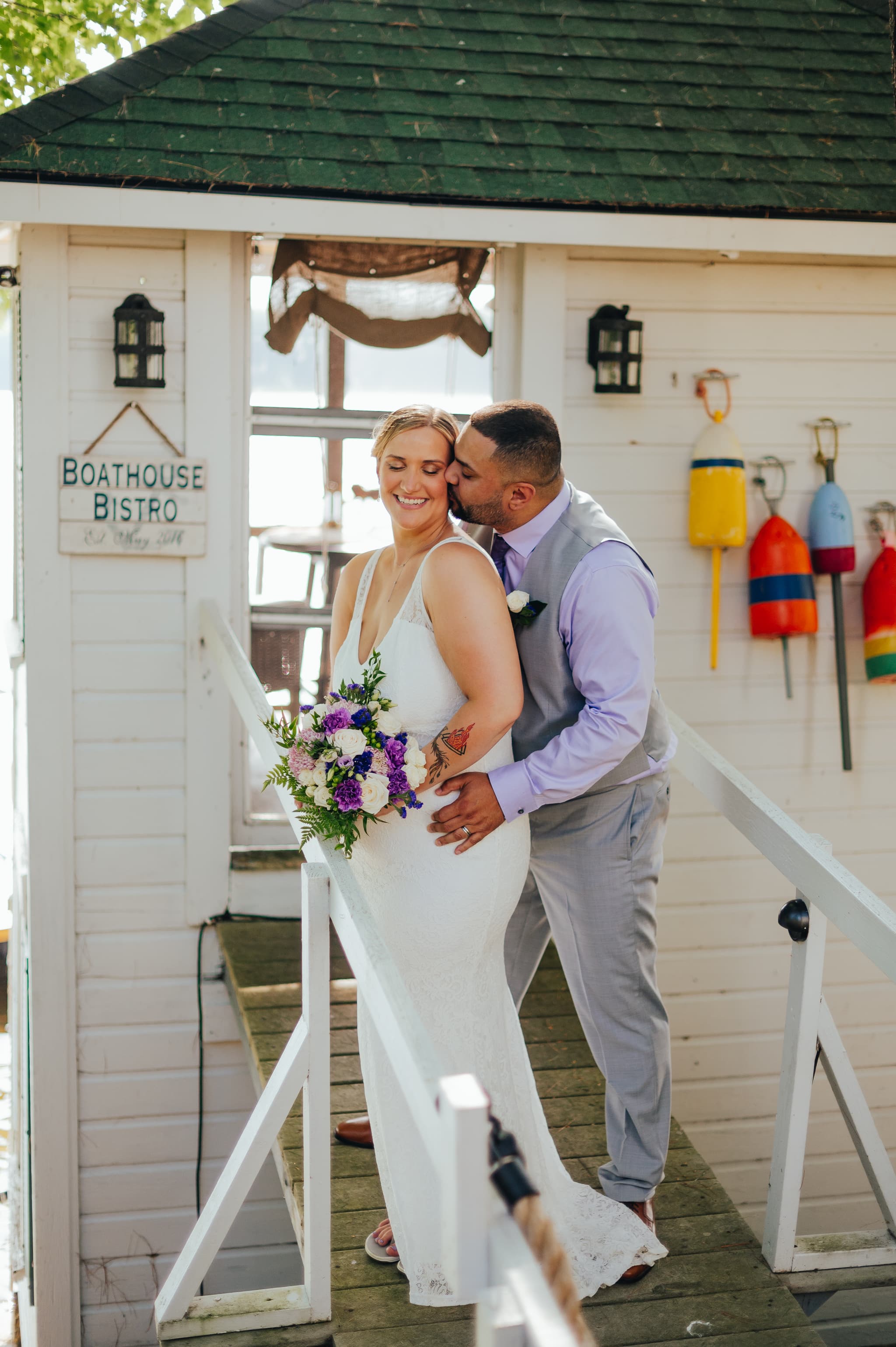 A happy couple embraces on a wooden dock in front of a charming boathouse, with the bride holding a bouquet of flowers.