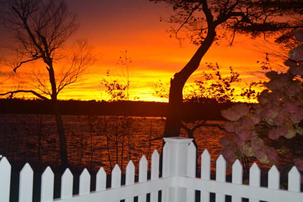 A vibrant sunset over a lake, framed by silhouetted trees and a white picket fence.