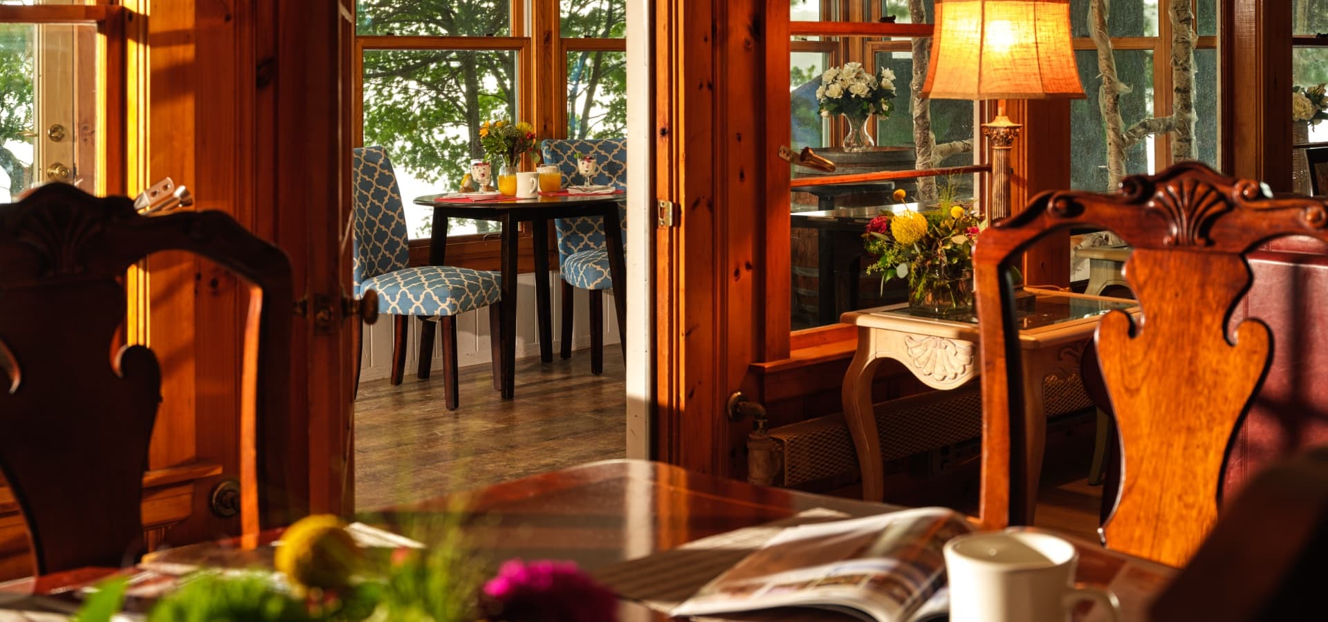 A cozy dining space with two blue-patterned chairs near a window, surrounded by wooden accents and a bright lamp.