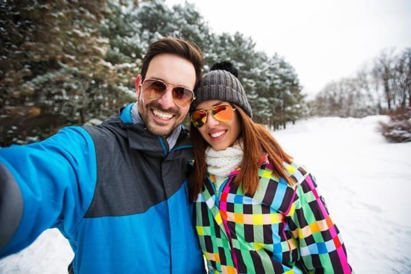 A smiling couple poses for a selfie in a snowy landscape, wearing colorful winter jackets and sunglasses.