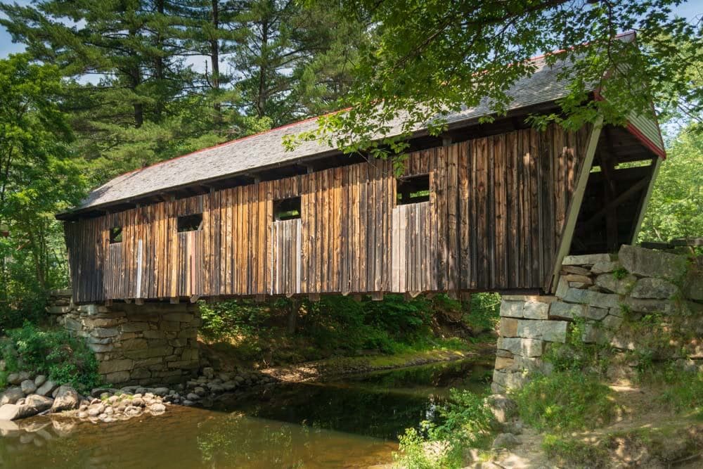 A wooden covered bridge spans a tranquil stream surrounded by lush greenery.