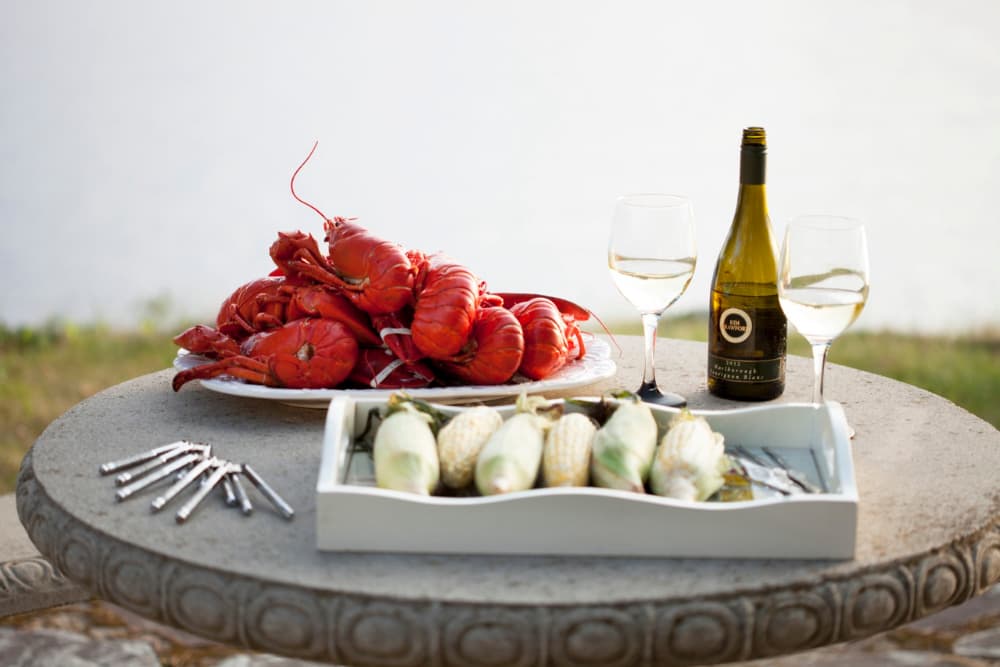 A plate of cooked lobsters, a tray of corn, two glasses of white wine, and a bottle of wine are arranged on a stone table by the water.