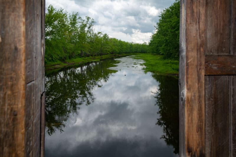 A serene river reflecting trees and clouds, viewed through an open wooden frame.