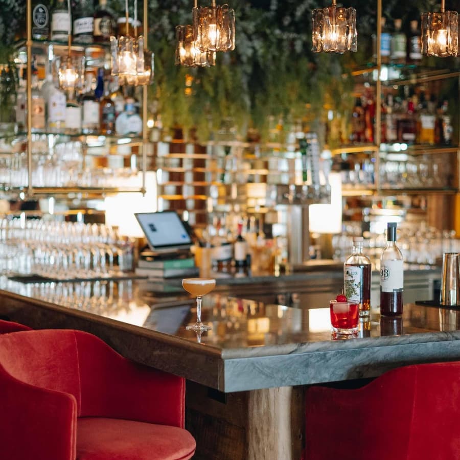 A stylish bar featuring red chairs, elegant lighting, and a selection of drinks on a marble countertop.