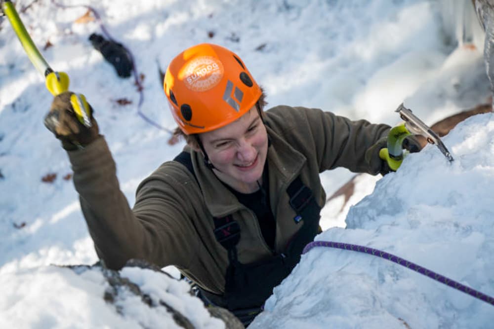 A climber smiles while ascending a snowy slope, equipped with ice axes and wearing a helmet.