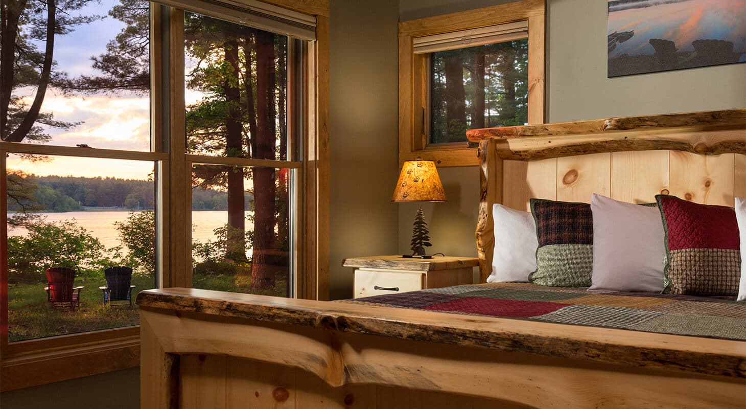 Cozy rustic bedroom with a wooden bed, overlooking a serene lake at sunset.