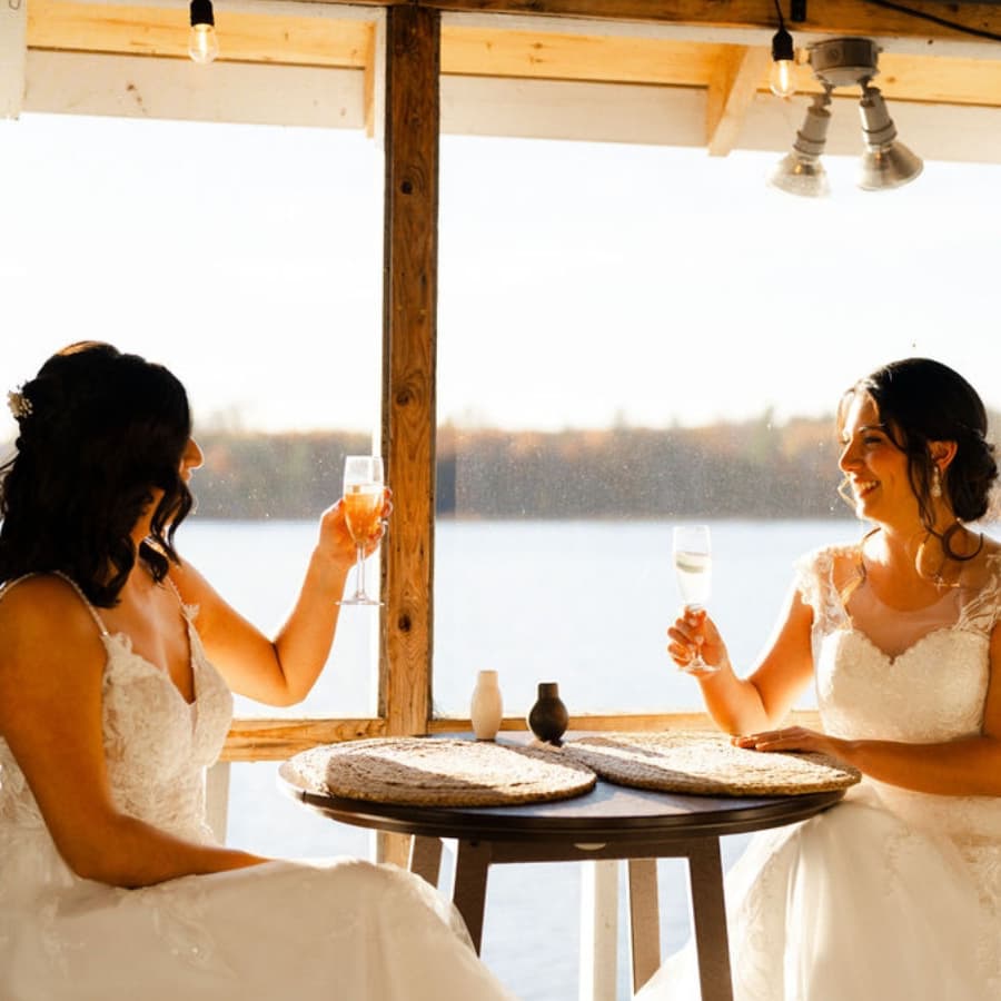 Two brides toast with champagne glasses at a lakeside table.