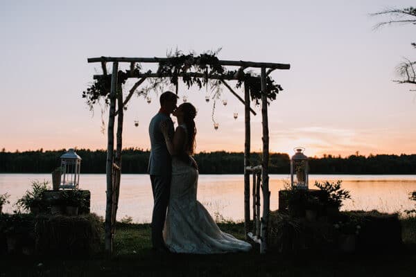 A couple kisses beneath a rustic arch at sunset by the water.