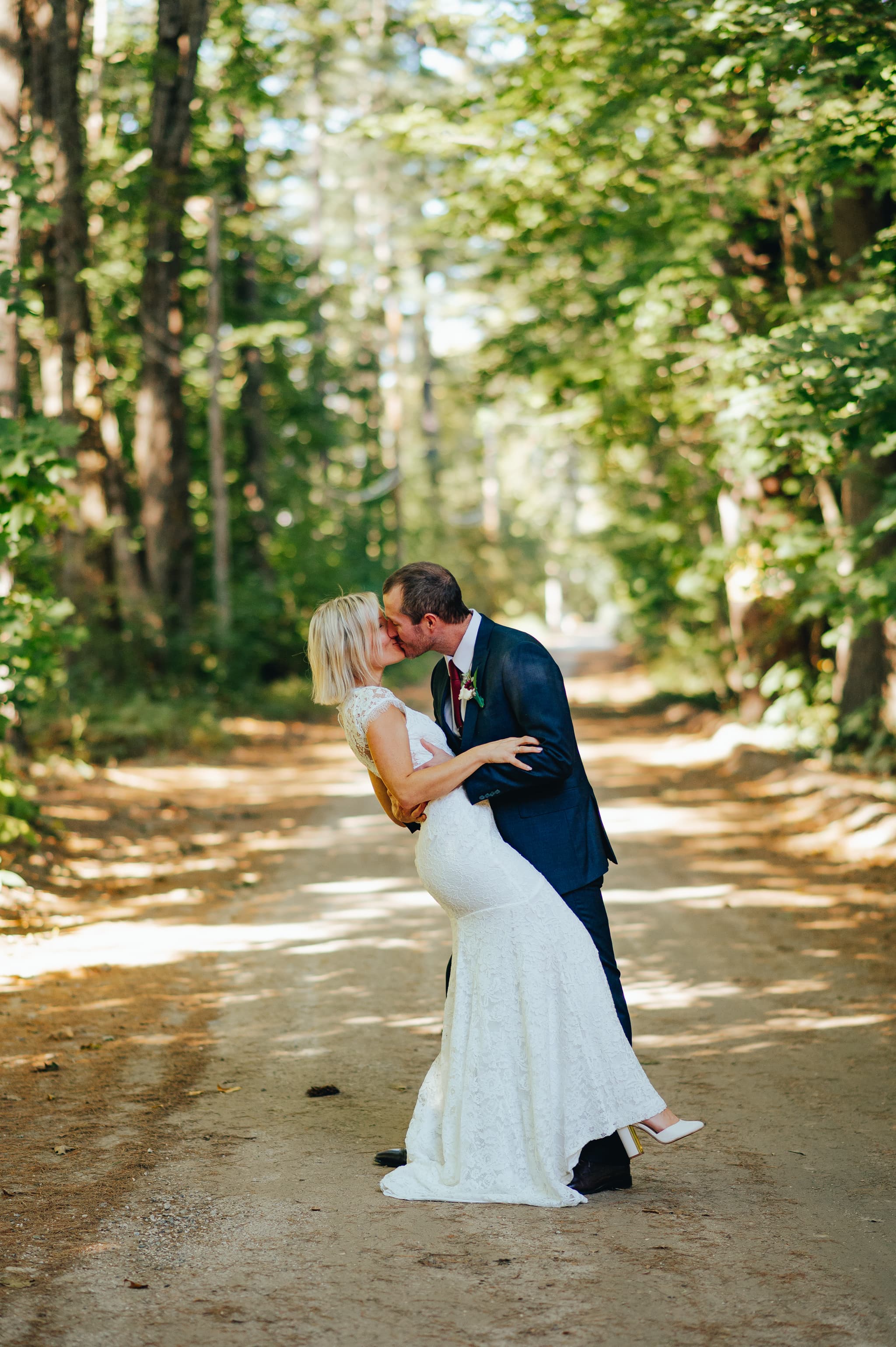 A couple shares a kiss on a sunlit dirt path surrounded by trees.