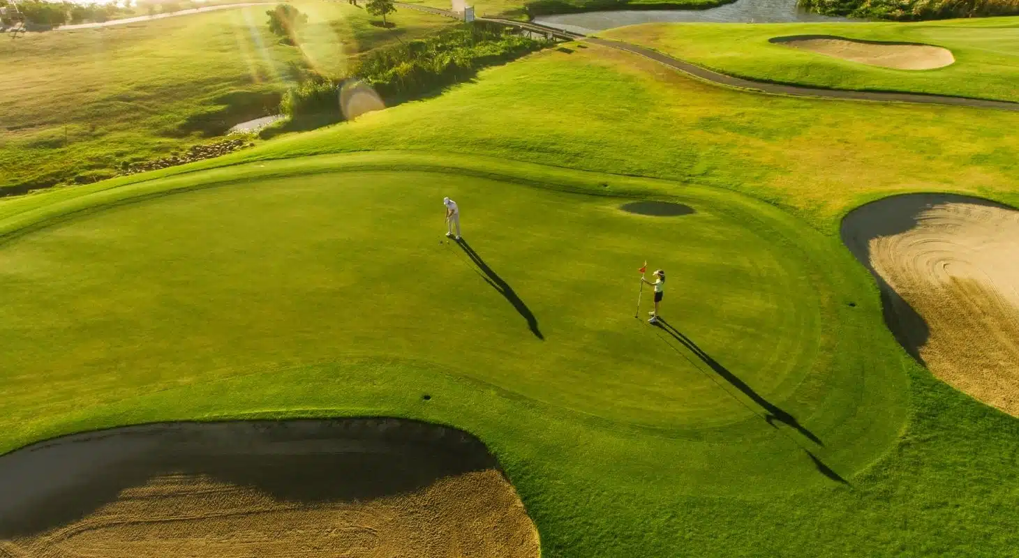 Two golfers on a lush green putting green surrounded by sand traps and water hazards.