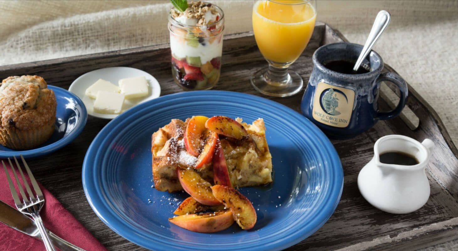 A wooden tray holds a blue plate of baked French toast topped with fruit, accompanied by a muffin, butter, a parfait, orange juice, and a coffee mug.
