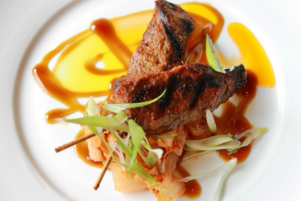 Grilled meat skewers served with kimchi and drizzled with sauce on a white plate.