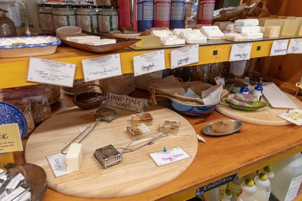 A wooden shelf displays various handmade soaps and natural products with labeled containers.