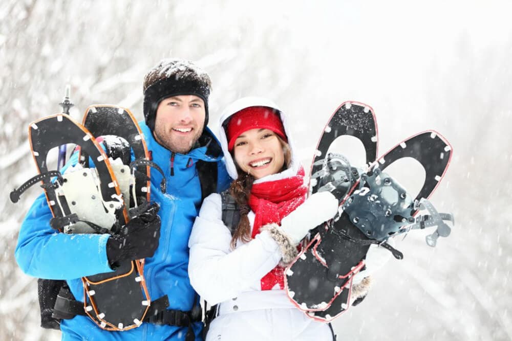 A smiling couple holds snowshoes while standing in a snowy landscape.