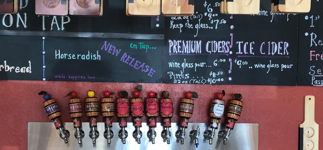 A wall display featuring multiple cider taps and a chalkboard menu with drink prices and descriptions.