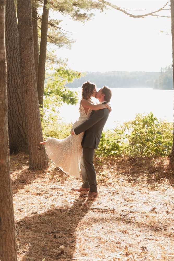A couple shares a kiss in a sunlit forest by a lake, with the bride lifted in the groom's arms.