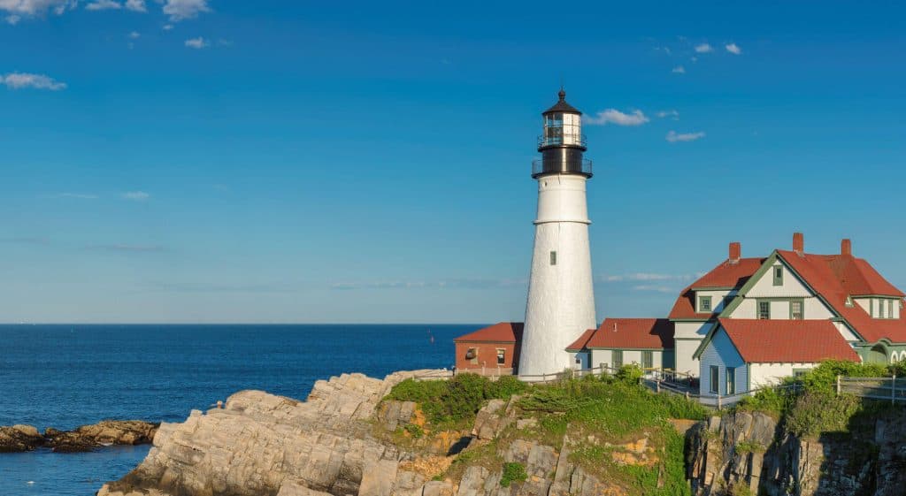 A white lighthouse stands on a rocky shore beside a red-roofed house, with the ocean and blue sky in the background.