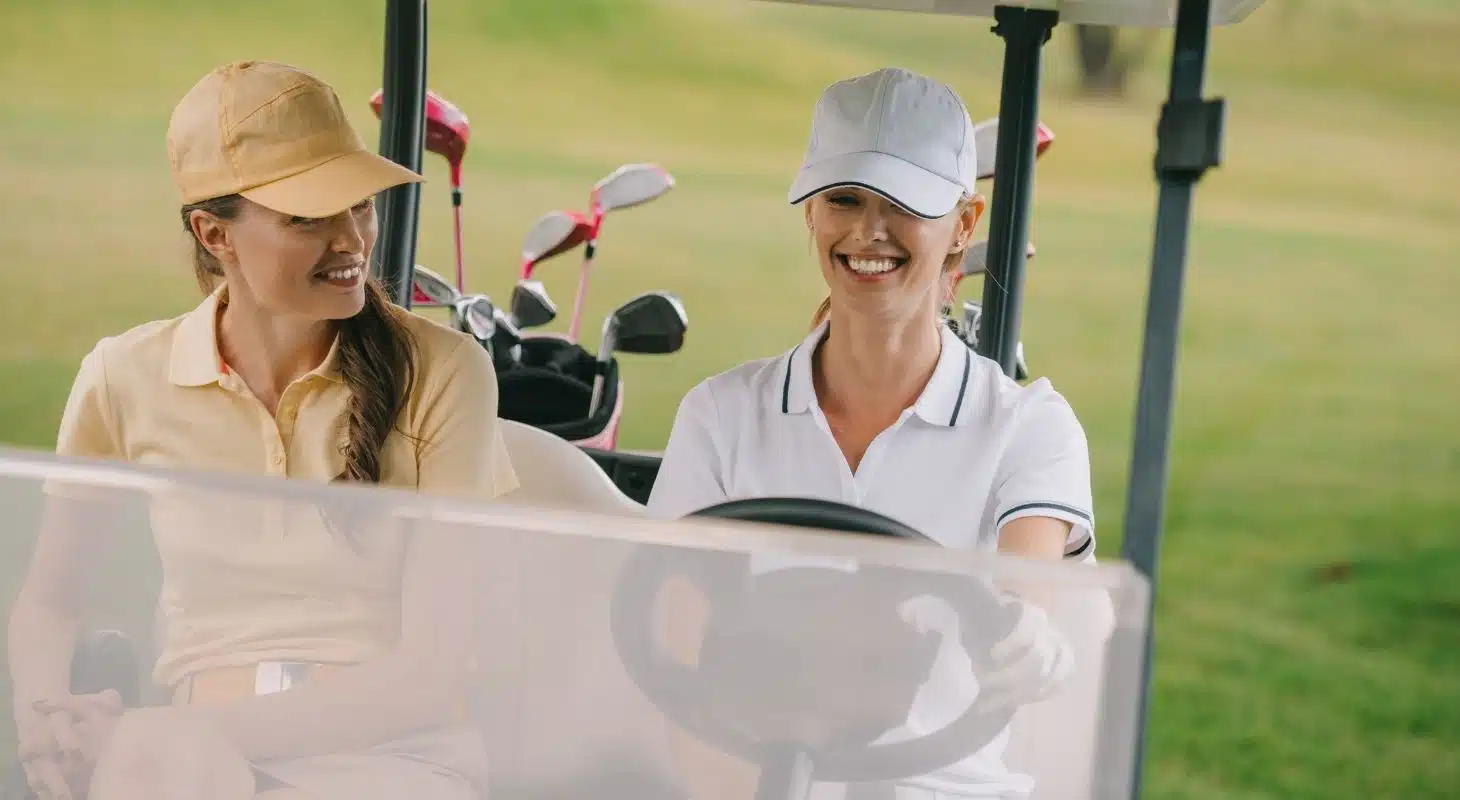 Two women in golf attire smiling and enjoying a ride in a golf cart. Two women in golf attire smiling and enjoying a ride in a golf cart.