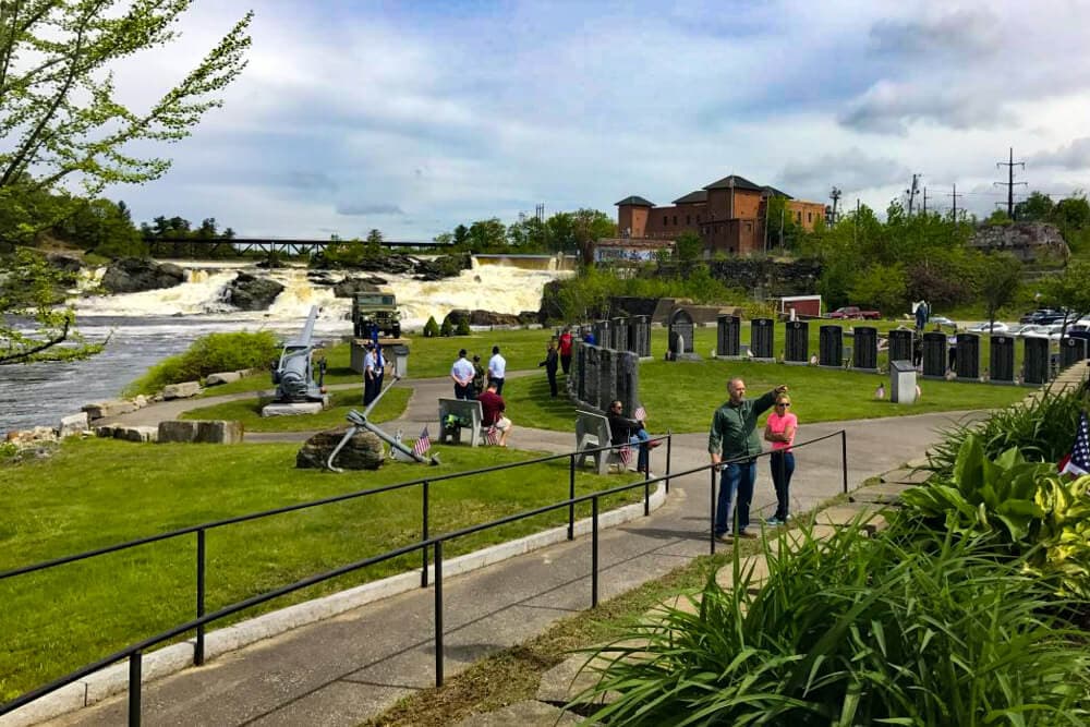 A group of people stroll along a path by the river, with a waterfall and historic buildings in the background.