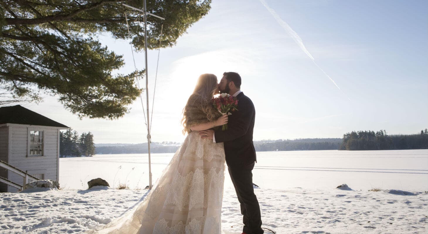 A bride and groom share a kiss on a snowy lakeside, framed by sunlight and trees.