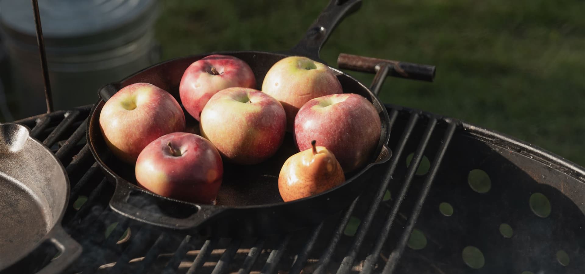A cast iron skillet on a grill holds several red apples and one small pear.