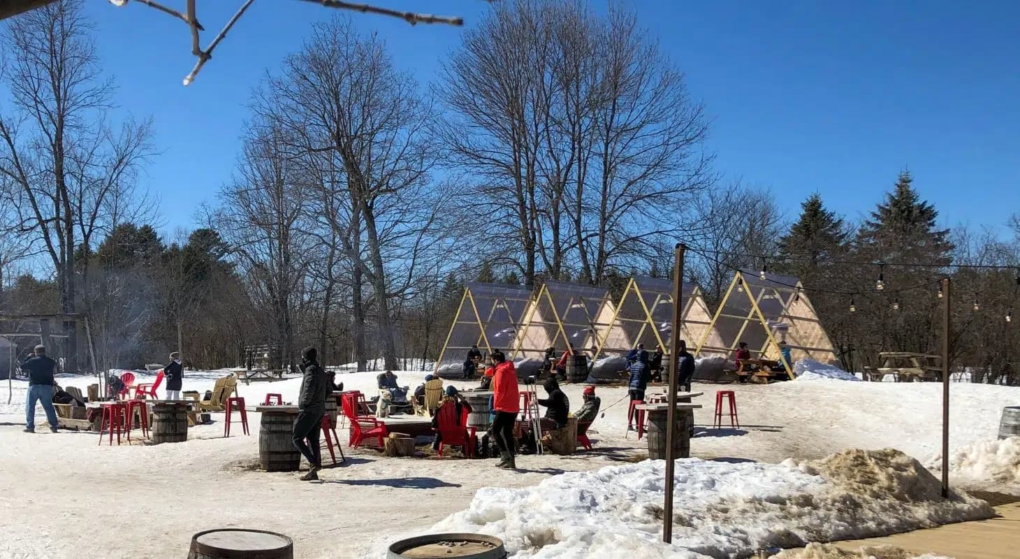 People gather around outdoor fire pits in a snowy landscape with tents and trees under a clear blue sky.
