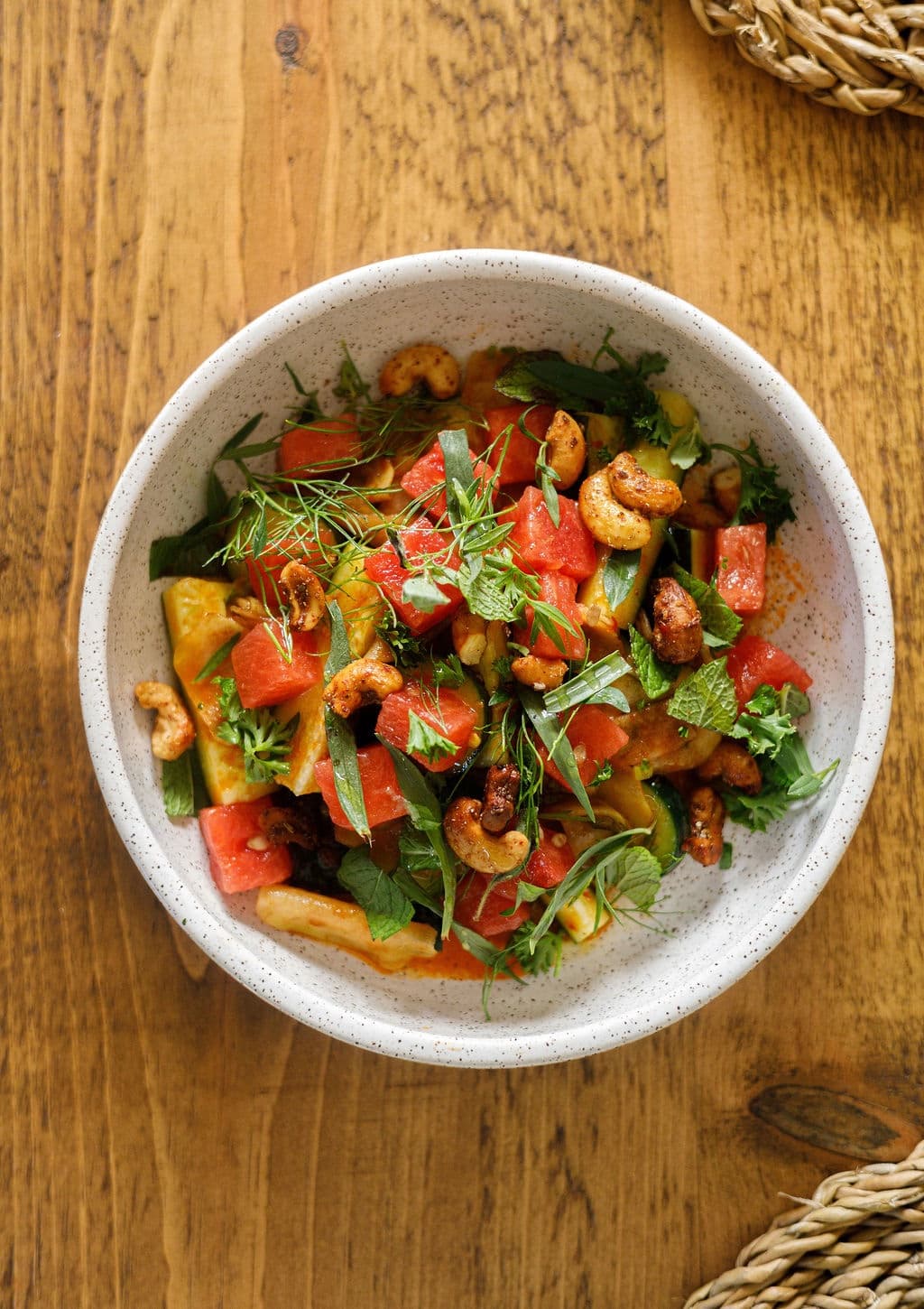 A vibrant salad with watermelon, herbs, and cashews served in a speckled bowl on a wooden surface.