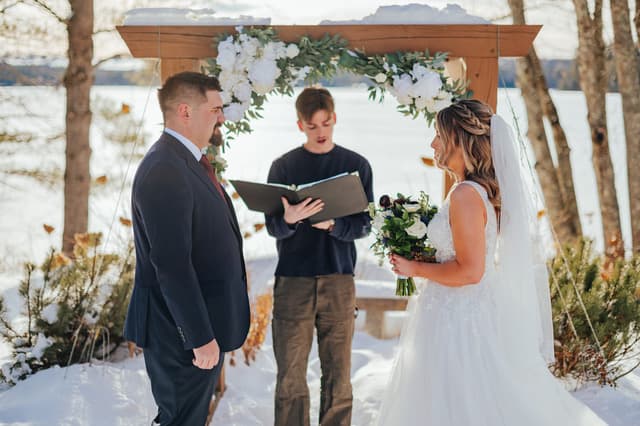 A couple stands under an arch adorned with flowers while a person reads from a book during their outdoor wedding ceremony in a snowy setting.