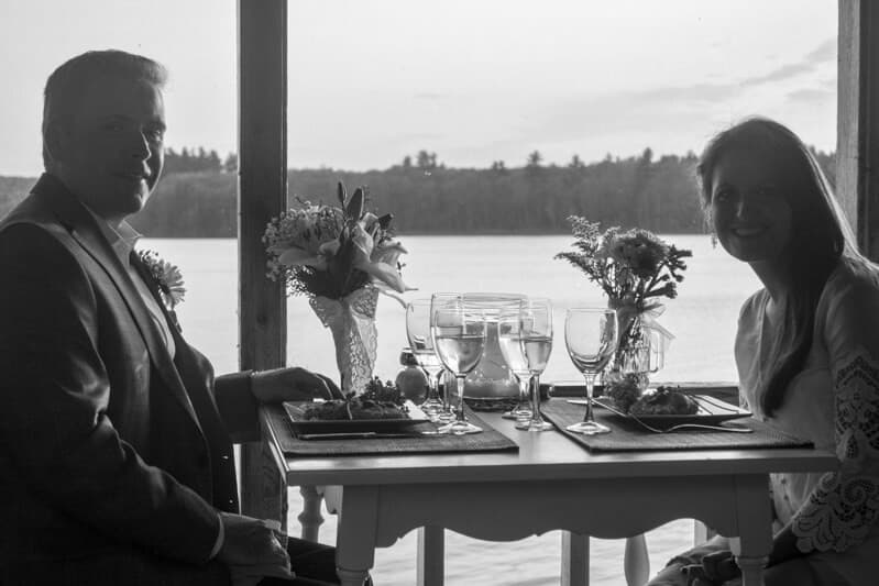 A couple sits at a table with flowers and dinnerware, enjoying a lakeside meal at sunset.