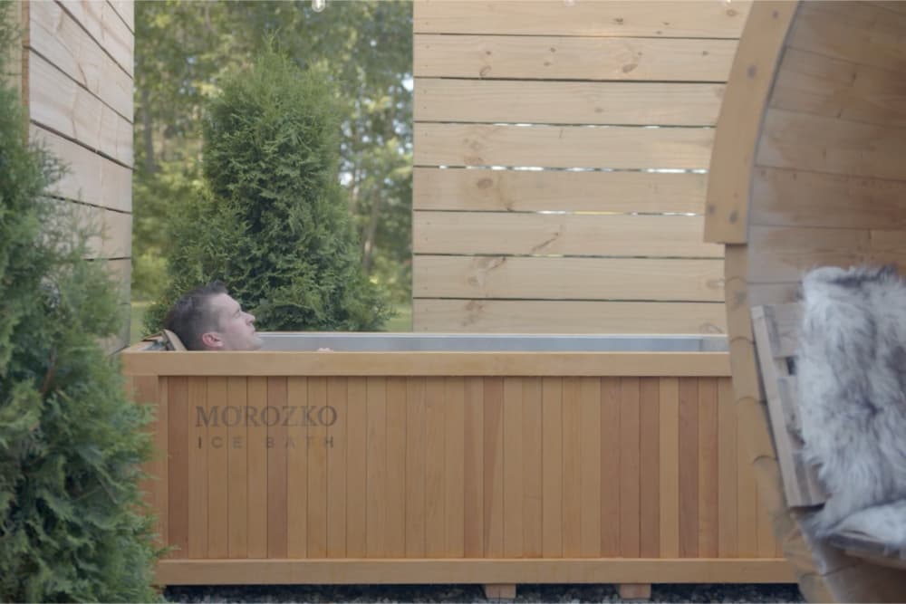 A person relaxes in a wooden ice bath surrounded by greenery.