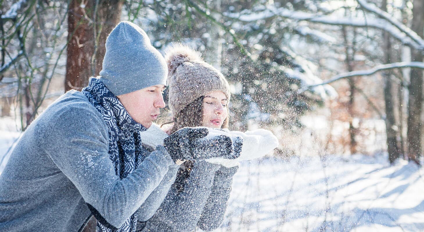 A man and woman in winter attire blow gently into the air, creating a flurry of snow in a snowy forest setting.