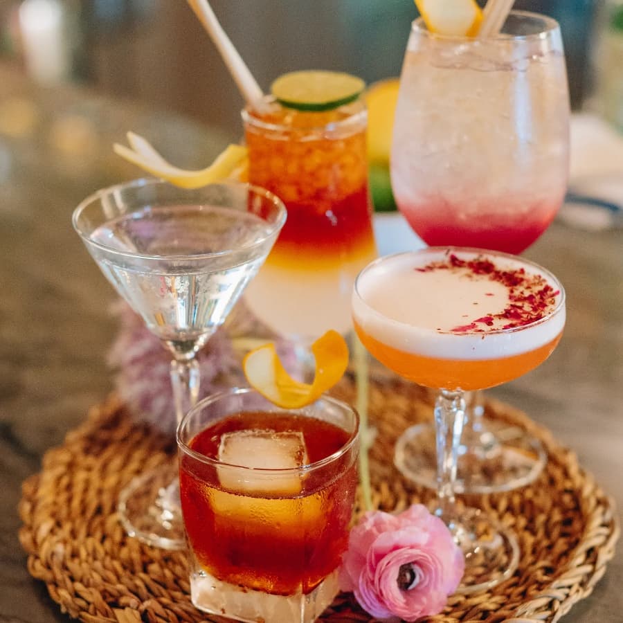 A variety of colorful cocktails served on a woven platter with a flower accent.
