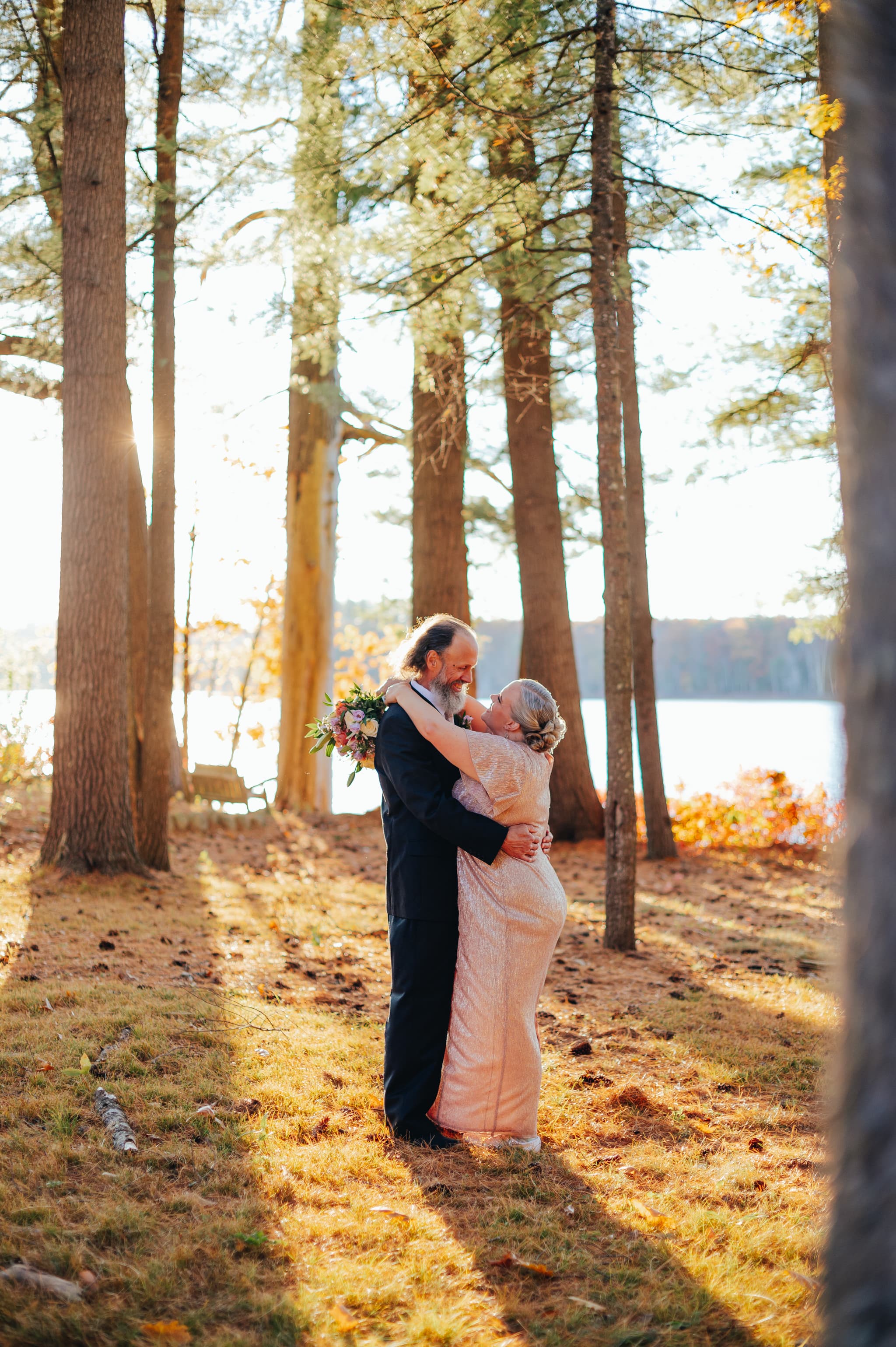 A couple embraces in a sunlit forest, celebrating their love against a backdrop of trees and a serene lake.