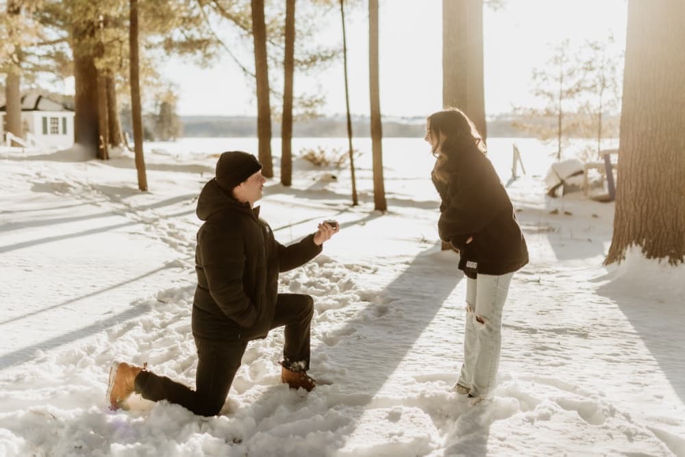 A man kneels in the snow to propose to a woman, who looks surprised and happy.