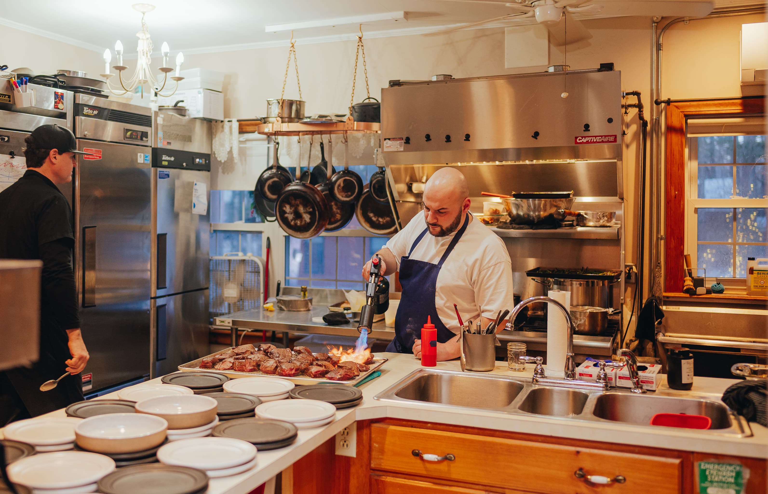 A chef uses a torch to sear meat in a bustling kitchen with various cooking tools and utensils.